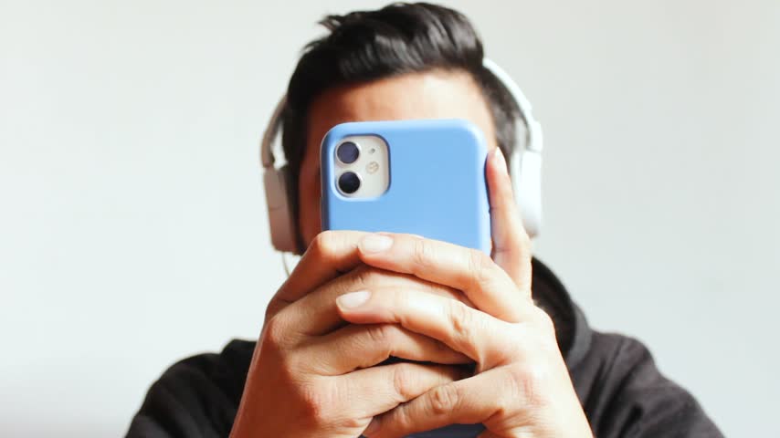 Portrait of a young Latino man chatting on a smartphone while listening to music with wired headphones, against a white background. Concept of lifestyle and technology