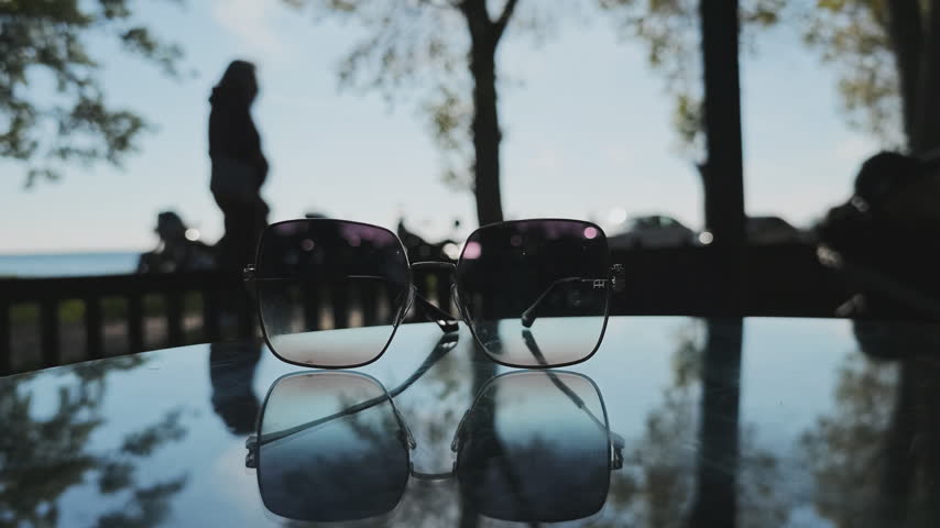 Sunglasses resting on a reflective glass table with people walking in a park and trees reflecting on the surface