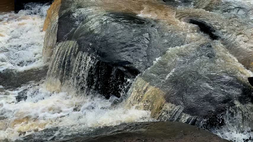 Closeup view of fast flowing stream water cascading over smooth dark rocks and creating foamy white rapids