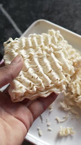 Hand holding a block of raw instant noodles over a white plate