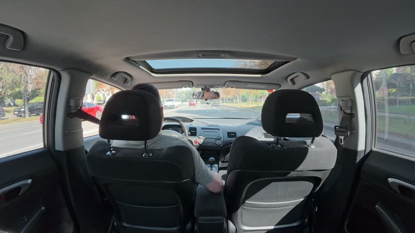 Static wide angle shot from rear passenger seat showing driver steering automobile down sunny urban avenue with traffic.