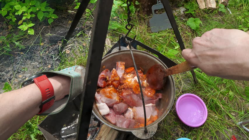 Point of view shot of male hands stirring raw pork meat with vegetables in a cauldron hanging over fire. Campsite cooking scene under natural daylight.