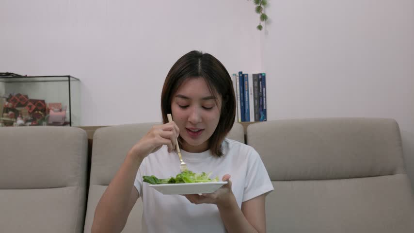 Young woman sitting at home, holding a plate of fresh salad and eating lettuce with a fork. Healthy eating, diet, wellness, and home lifestyle concept.