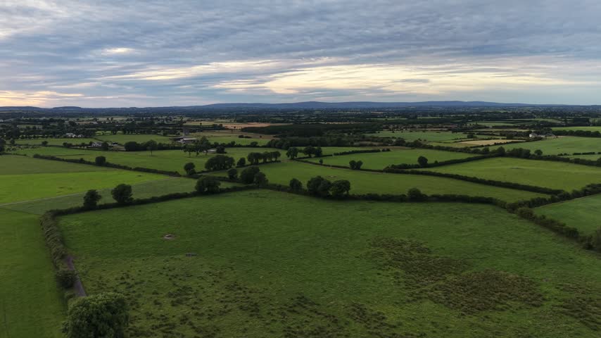 4K aerial view of green agricultural fields divided by hedgerows beneath a cloudy sky with a distant rural horizon.