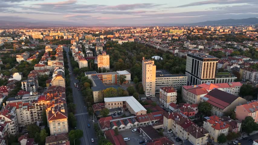 Aerial drone view over central Sofia, Bulgaria, showing dense urban neighborhoods, residential buildings, tree lined streets, red rooftops and modern architecture illuminated by warm evening sunlight.