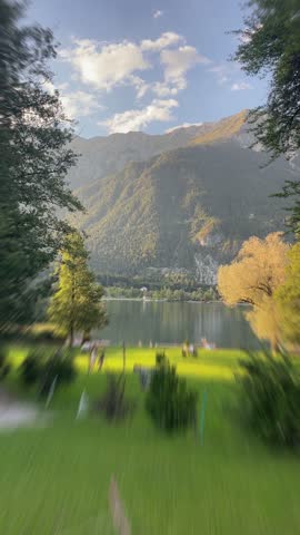 Vertical lakefront motion toward grassy park shoreline framed by trees, reflective water, and sunlit forested mountain ridge in warm late afternoon light, scenic