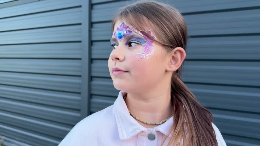 Young girl with colorful face paint looks into distance then toward camera, expressive child portrait