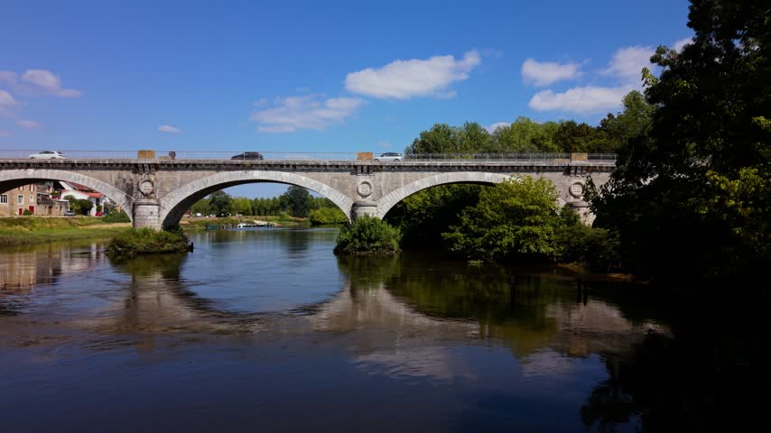 Aerial view pan left of a countryside bridge in France with a car moving along it beside a river