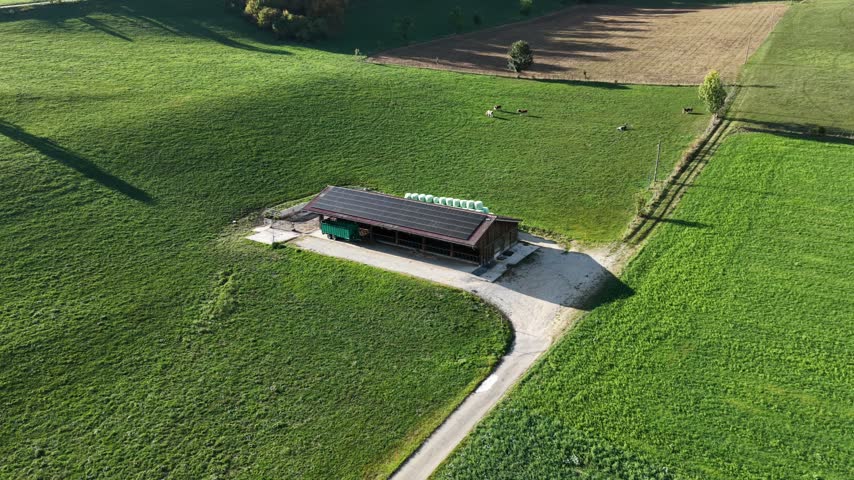 Green fields surround a simple barn structure. Clear skies cast soft shadows across the landscape. Winding path leads to the barn entrance. Lush countryside offers peaceful, rural imagery
