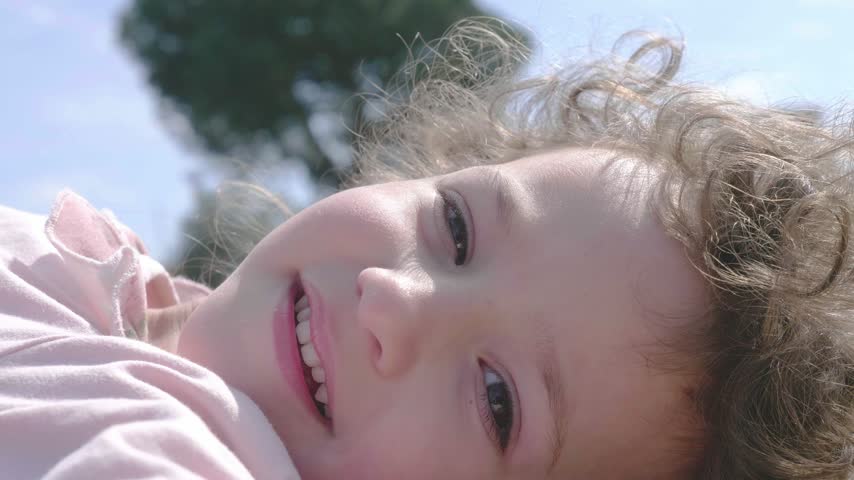 A young girl with curly hair and a pink sweater shares a serene smile with a camera to her left, her expression reflecting the tranquil joy of a sunny garden day