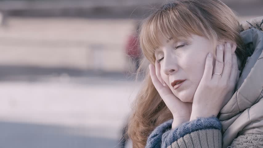 Amidst the backdrop of a historic public square, a middle-aged woman with red hair holds her face in her hands, her gaze forward yet lost in deep thought