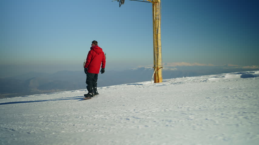 A man wearing a red jacket gracefully snowboards down a pristine snowy mountain slope. He enjoys the thrilling winter sport amidst the clear blue sky and picturesque mountain landscape.