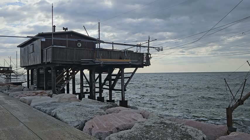 Wooden Fishing Hut on Stilts Over Rocky Coast With Nets and Cloudy Sea Sky