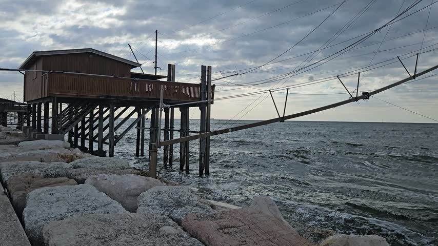 Wooden Fishing Hut on Stilts Over Rocky Coast With Nets and Cloudy Sea Sky