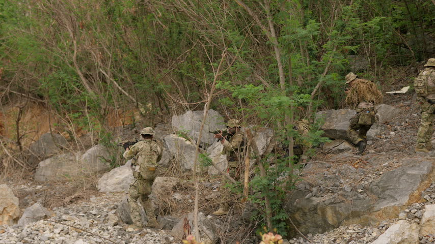 A team of soldiers in full combat gear conducts a tactical patrol, moving cautiously with rifles through a rugged, rocky landscape during a military operation.