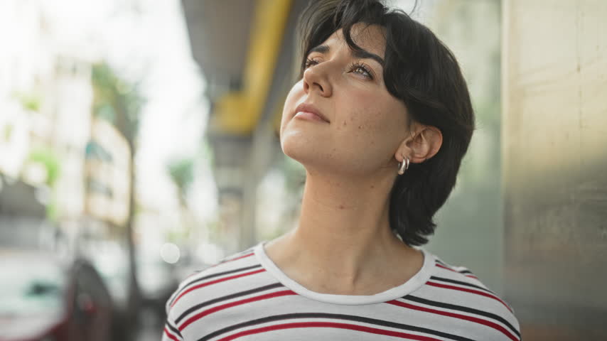 Young woman looks up, face and hoop earring visible on a street with glass storefronts, wearing a striped shirt; serenity reflection.
