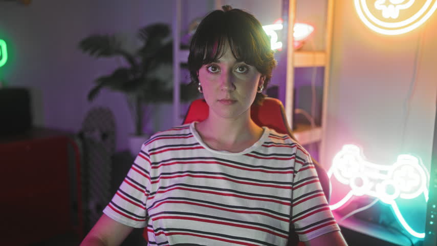Young woman with short hair sits in gaming chair arms crossed under neon controller sign in studio; serious concentration.