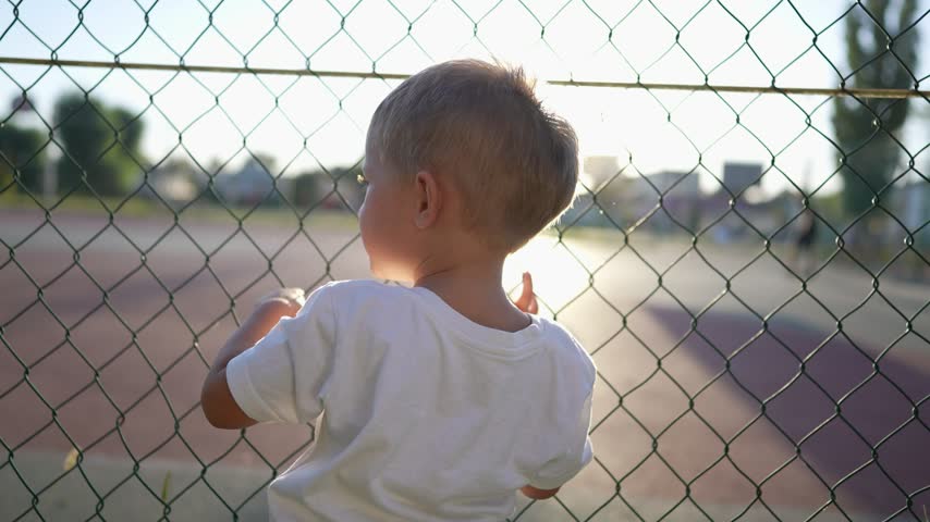 boy stands near the playground. concept of happy childhood and loving family. a child holds on to the net of a sports ground, court and glare of the sun in the background lifestyle