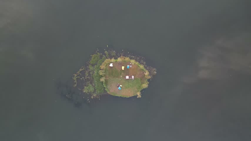 High-angle drone shot capturing a tiny grassy island with colorful camping tents surrounded by calm, dark lake water, emphasizing solitude and nature adventure.