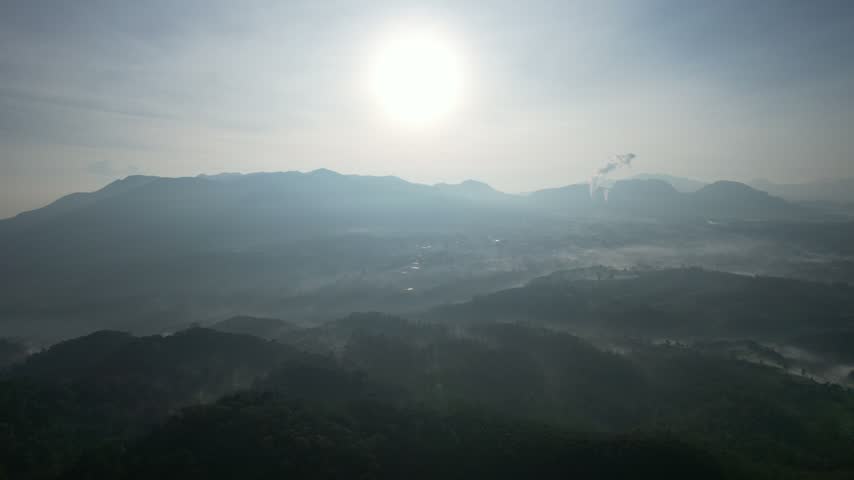 Breathtaking aerial footage of a misty mountain range at dawn. The scene captures layers of hills shrouded in fog under a bright, hazy sun with distant industrial smoke plumes.