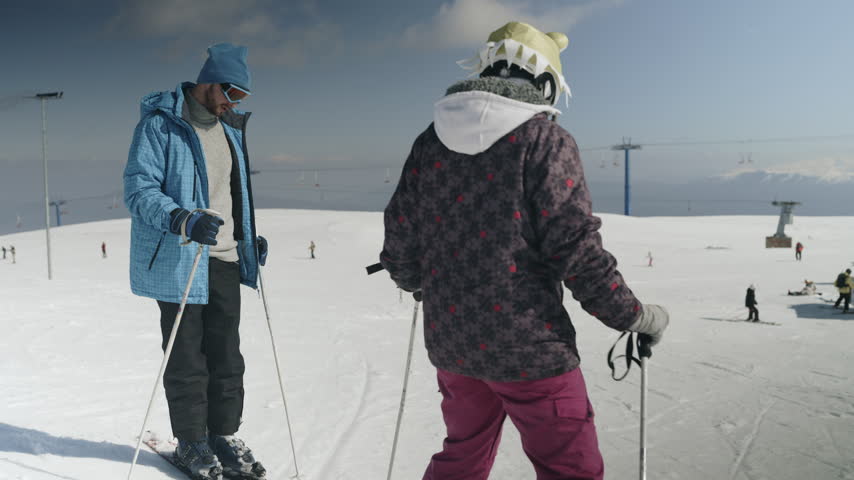 A man and a child are skiing down a snowy mountain slope. They enjoy winter sports at a busy ski resort on a bright day.