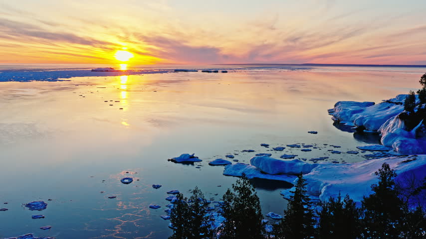 Slow aerial drift follows the icy Presque Isle Park shoreline as warm sunrise light reflects across calm Lake Superior in Marquette, Michigan.