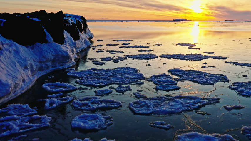 Drone pullback reveals floating shoreline ice, calm open water and a bright winter sunrise over Lake Superior at Presque Isle Park.