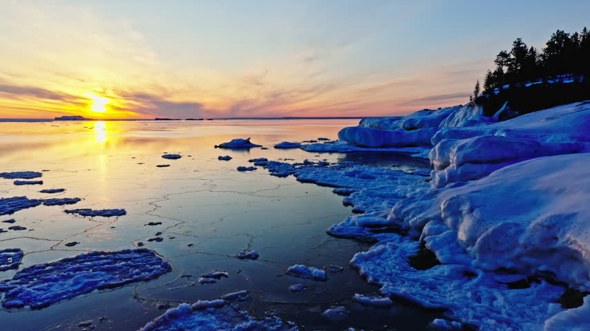 Lateral drone move follows a sculpted ice-covered shoreline at Presque Isle Park while the sun rises over calm Lake Superior in Marquette, Michigan.