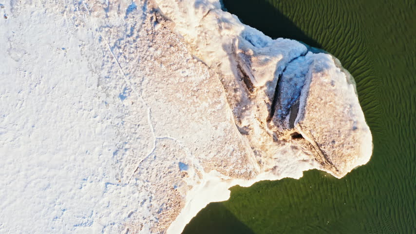 Top-down aerial travels over a snow-covered Lake Superior shoreline with sculpted ice shelves and dark open water at Presque Isle Park.