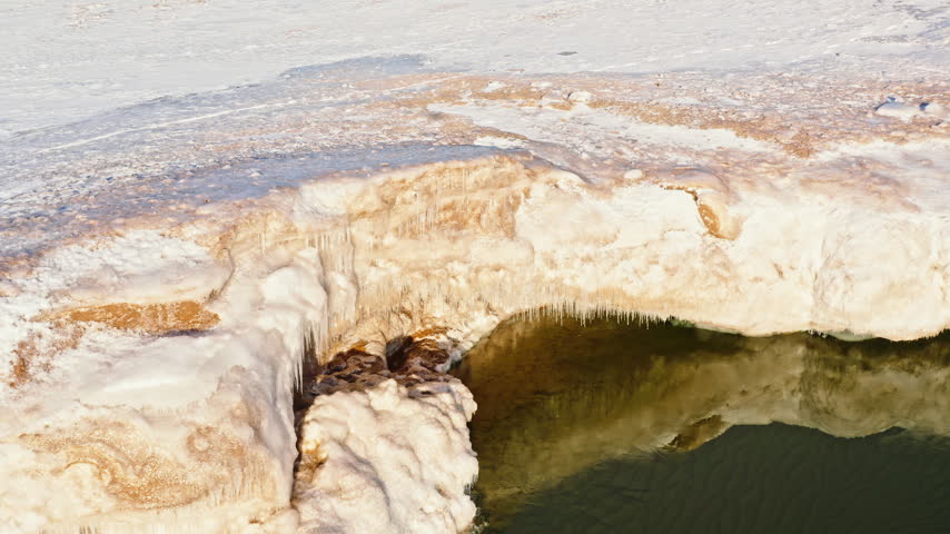 Top-down aerial looks straight down on a thick sculpted ice shelf, snow-covered shoreline and dark Lake Superior water at Presque Isle Park.