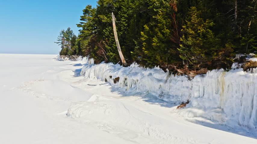 Low aerial tracking view follows a snow-covered Lake Superior shoreline lined with evergreen forest, frozen cliffs and hanging ice formations in Michigan
