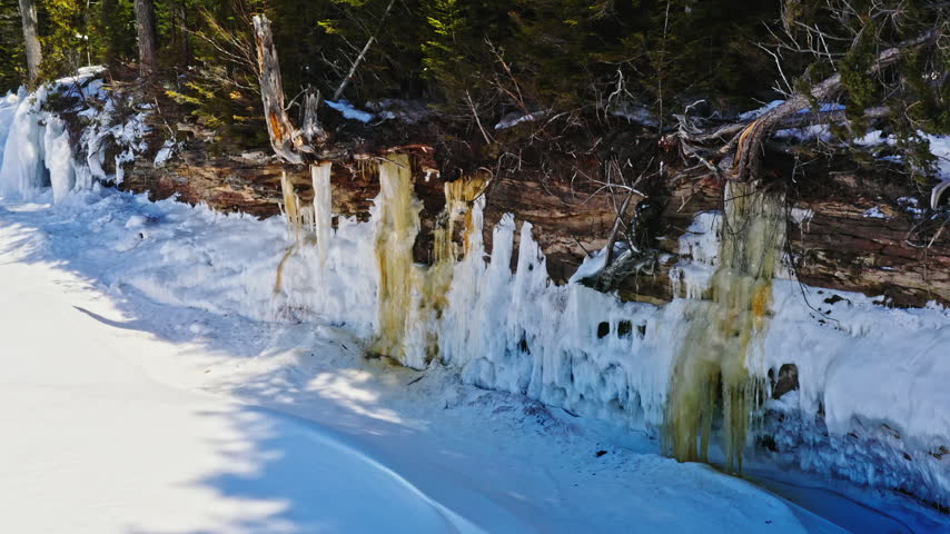 Low drone pass moves beside tall hanging icicles and layered ice formations along a snowy forested shoreline in Michigan