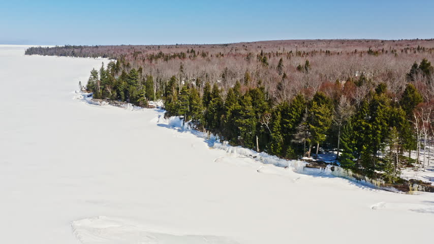 High aerial view looks across a broad frozen lake or bay beside a snow-covered evergreen and hardwood forest in Michigan