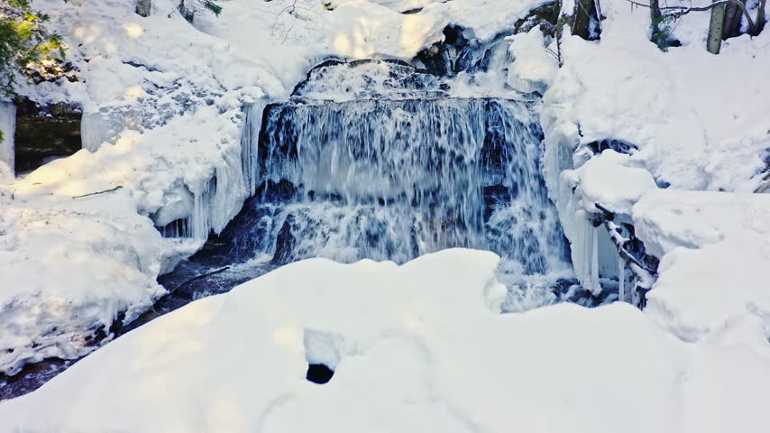 Ground-level winter view shows a small broad waterfall flowing behind a deep rounded snow bank with ice and evergreen forest surrounding the creek.