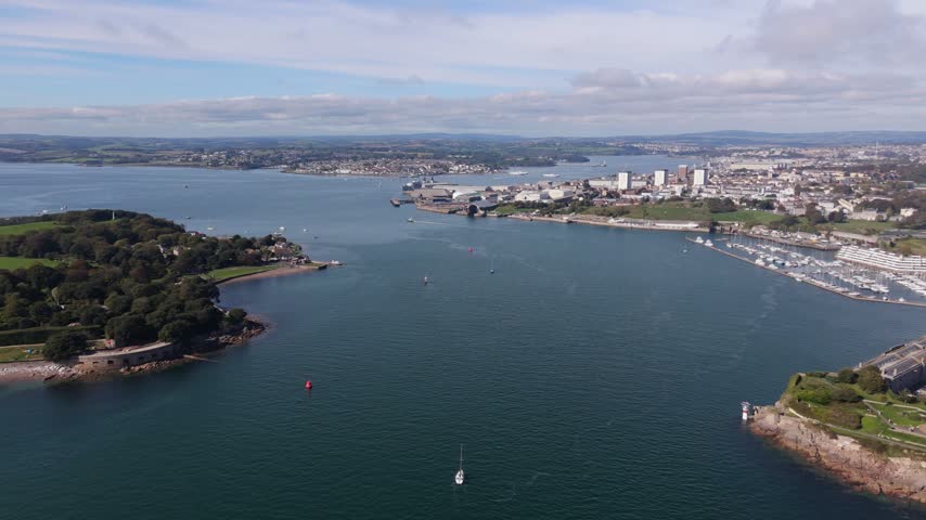 An expansive aerial panoramic view showcases the city of Plymouth, its busy harbor filled with yachts, and the historical fortifications along the coastline, all under a partly cloudy sky.