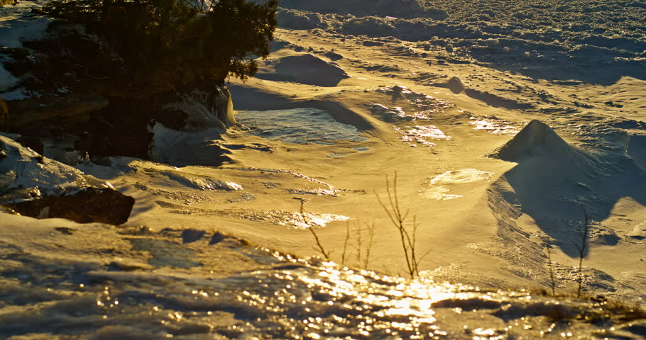 Camera tilts upward from wind-shaped snow and shoreline trees to reveal a warm golden winter sunset over frozen Lake Superior near Marquette, Michigan.