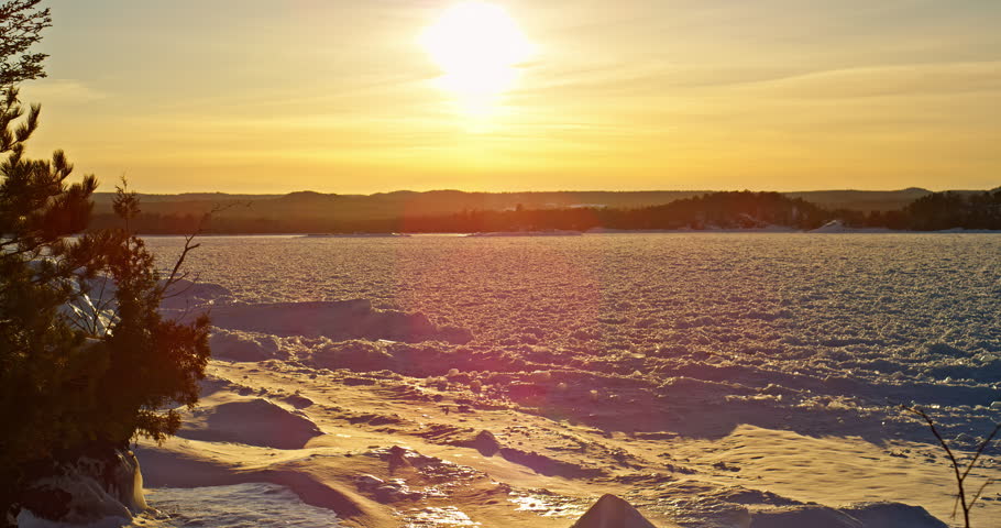 Slow leftward pan follows a golden winter sunset across frozen Lake Superior with snow-covered shoreline and dark trees in the foreground.