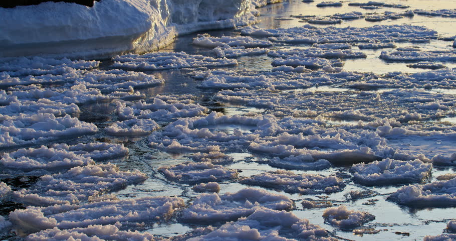 Camera tilts upward from floating Lake Superior pancake ice to reveal a dark basalt shoreline coated with thick snow and ice at Presque Isle Park.