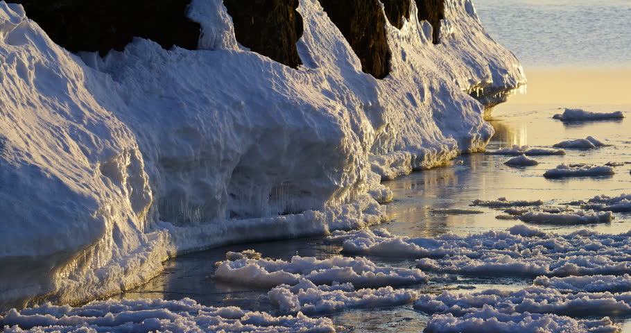 Static close winter view shows thick snow and golden-lit ice coating dark basalt shoreline beside floating Lake Superior pancake ice at Presque Isle Park.
