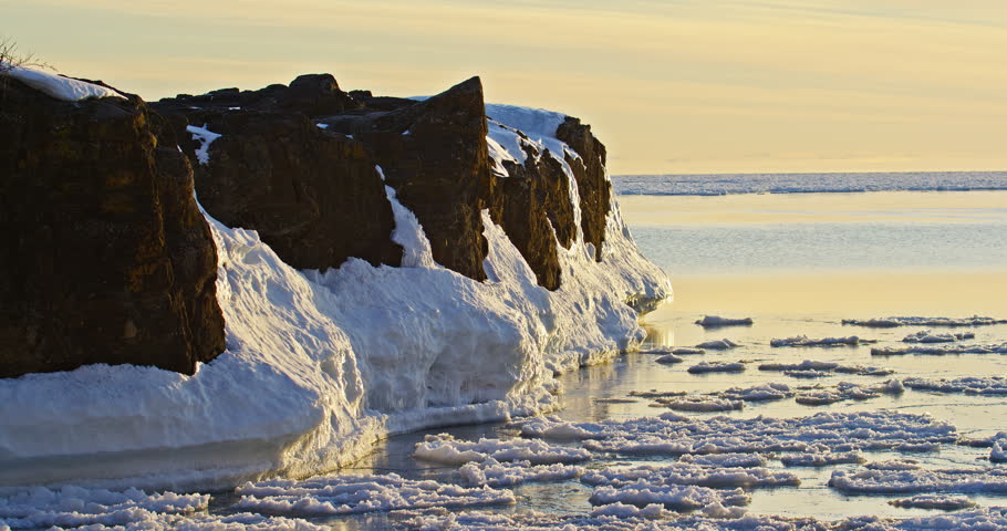 Static winter view shows dark basalt Black Rocks coated with snow and ice beside Lake Superior pancake ice in warm sunset light at Presque Isle Park in Marquette, Michigan.