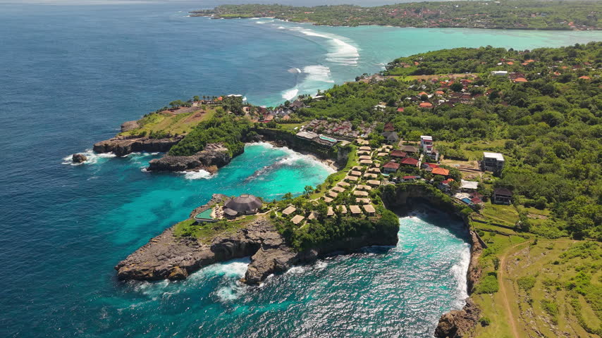 Waves crashing into a rocky cove surrounded by island vegetation and small resort huts along the coastline under bright sunlight