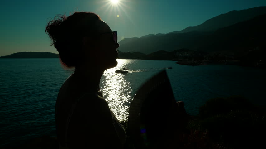 Silhouette of a woman with a fan enjoying the seaside sunset. Backlit silhouette of a woman with sunglasses and her hair in a bun fanning herself with a hand fan, enjoying the sea and the sunset