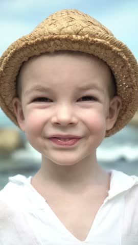 Cute young child wearing a woven sun hat smiles by the sea, joyful outdoor summer vacation.