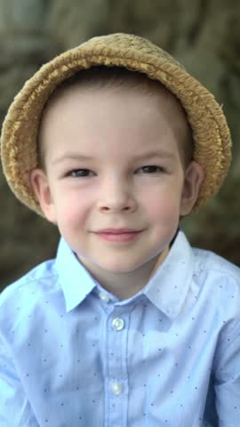 Smiling young boy in a straw hat and blue dotted shirt looks happily at the camera outdoors.