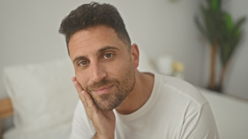 Man with hand on chin touching jaw while seated on bed inside a bedroom building, casual white shirt and subtle smile; contemplation.