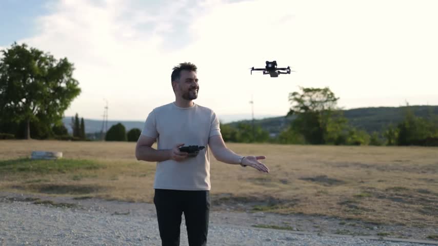 A man stands in an outdoor field, flying a drone and smiling. He uses a controller to guide the drone.