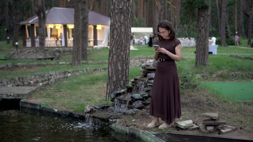 Young woman standing by an artificial waterfall with a phone. Girl wearing glasses and a dress stands beside a decorative waterfall with a festive tent in background.