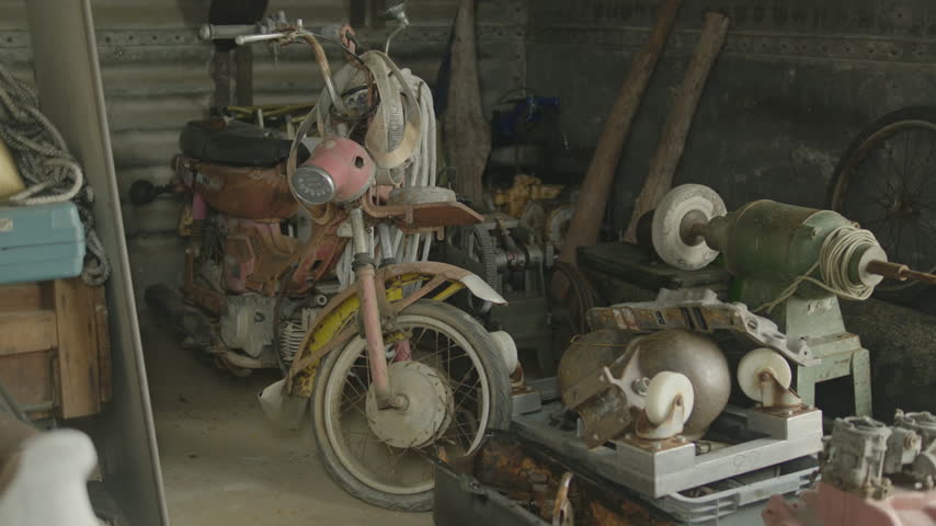 A rusty pink scooter sits among piled spare parts, a bench grinder, coiled ropes and other old tools inside a dim, cluttered shed