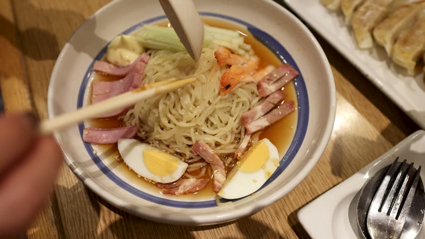 Top-down view of hands using chopsticks to eat cold ramen noodles in a restaurant bowl.