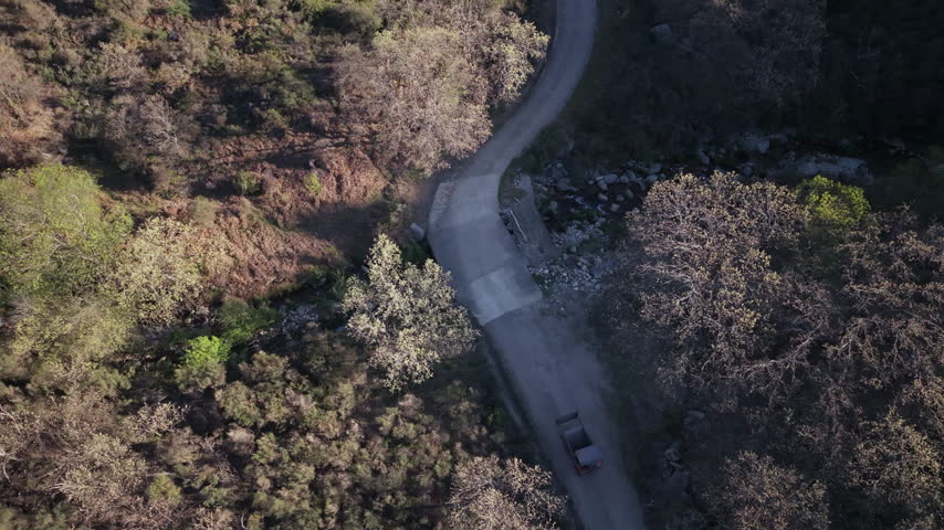 Top-Down Aerial Following Excavator Along Winding Dirt Road Bend, Rural Spain - Acebo, Sierra de Gata, Extremadura, Spain. Top-down drone shot that follows an excavator along a curving dirt road, tracking each bend. The road cuts through rocky shrubland and woodland, with long shadows and warm sunlight. 